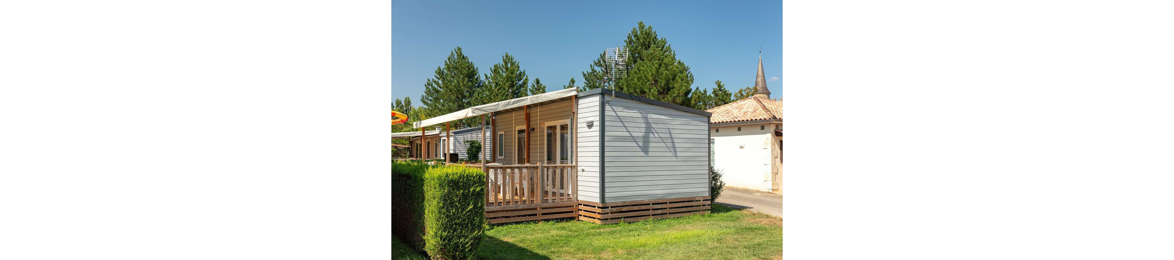 A modern mobile home with a wooden terrace stands in a sunny campsite. The structure features light-colored siding, large windows, and a covered porch area. Neatly trimmed hedges and green grass surround the unit, with other similar cabins visible in the background. Tall trees and a clear blue sky create a relaxed vacation atmosphere, while a nearby building and church steeple hint at a village setting.
