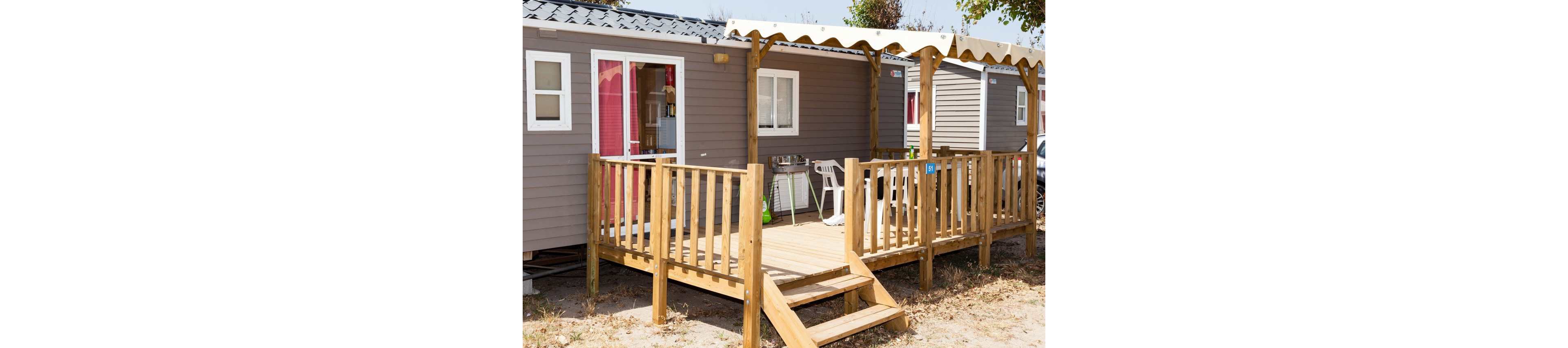 A modern mobile home with a wooden deck is shown in a sunny outdoor setting. The structure features gray siding, white-framed windows, and a glass door leading to the terrace. A wooden railing and small staircase provide access to the deck, which is partially shaded by a light-colored canopy. Outdoor chairs and household items are visible inside and on the deck, suggesting a relaxed vacation or campsite atmosphere.