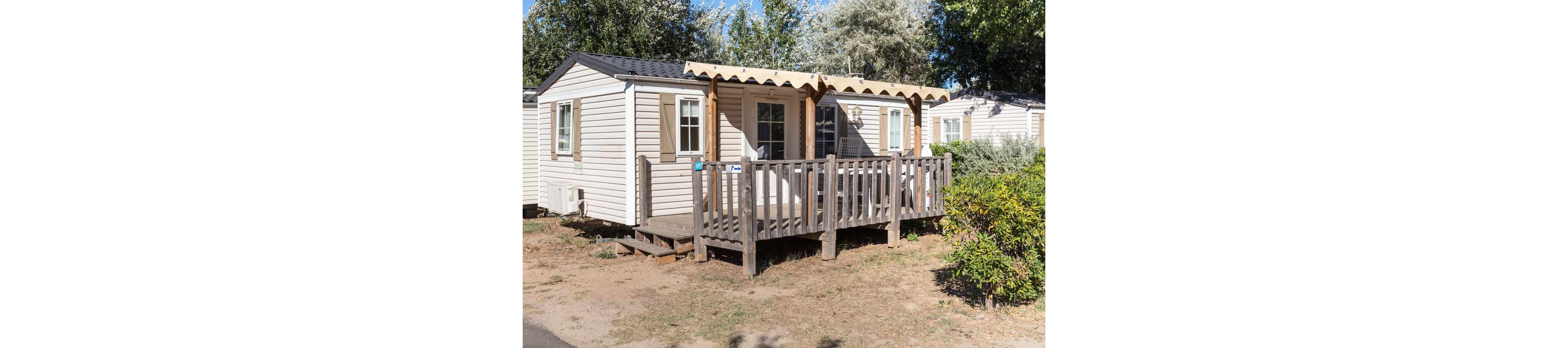 A compact mobile home with a wooden terrace stands in a sunny outdoor setting. The structure features light-colored siding, brown shutters, and a small covered porch with railing. Another similar unit is visible in the background, suggesting a campground or holiday park. Surrounding trees and shrubs provide greenery and shade around the homes.