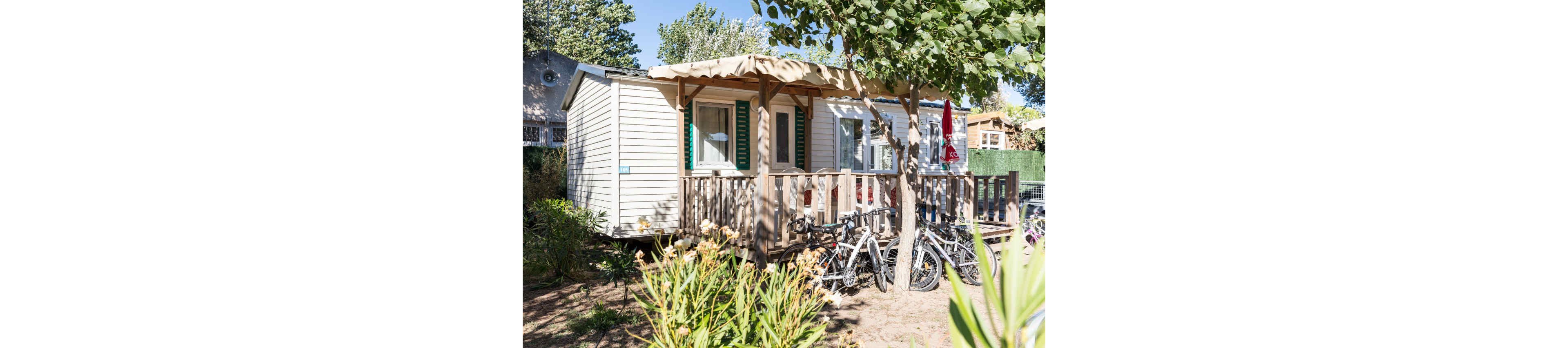 A small mobile home with a wooden deck is shown in a sunny outdoor setting. Several bicycles are parked in front of the porch, suggesting guests or family on a cycling holiday. Green trees and shrubs surround the structure, providing shade and a natural atmosphere. Bright daylight and clear skies create a relaxed, summery camping vibe.