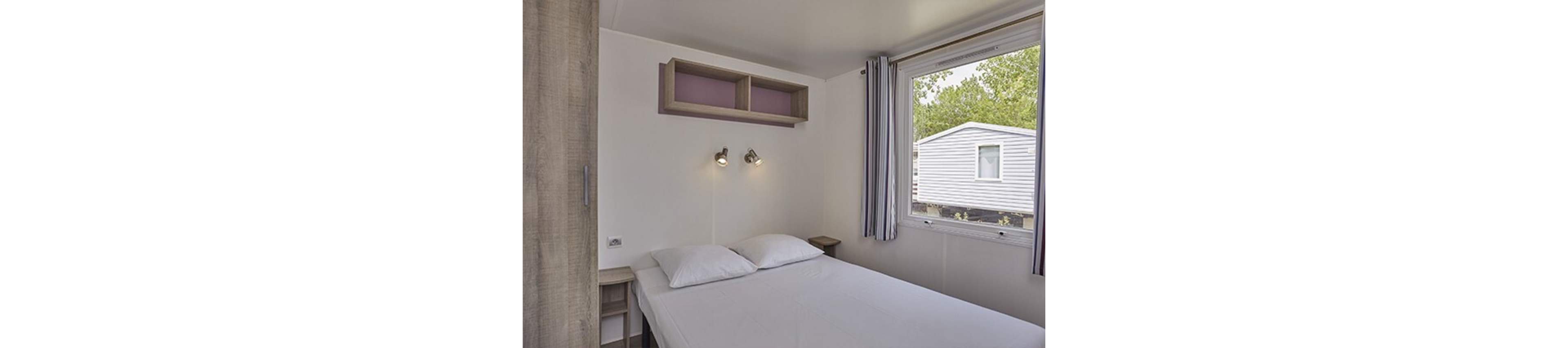 A small bedroom features a neatly made double bed with two white pillows against a plain wall. Two adjustable wall-mounted lights are positioned above the headboard, and open shelves provide simple storage. A large window with striped curtains lets in natural light and offers a view of a nearby mobile home or caravan and green trees outside. The overall decor is minimal and functional, creating a clean and cozy atmosphere.