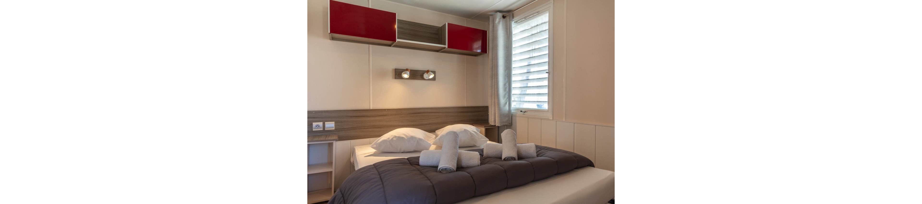 A compact modern bedroom features a double bed neatly made with a dark quilt and white pillows. Several rolled towels are placed on the bed, suggesting a hotel or guest accommodation. Above the bed, a wooden headboard and red wall cabinets add a contemporary touch. Natural light enters through a window with blinds and a light curtain, while two wall-mounted spotlights provide additional illumination.