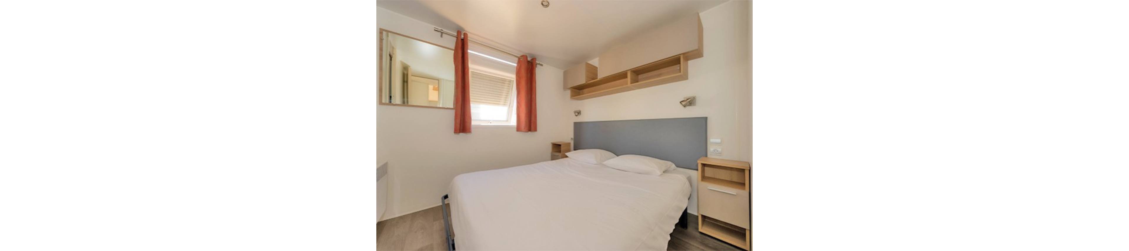 A compact bedroom features a neatly made double bed with a light gray headboard and white bedding. The room includes two small wooden bedside tables and built-in wall shelves above the bed. A window with partially closed blinds and orange curtains lets in natural light, reflecting in a large wall mirror. The neutral color palette and simple furnishings create a clean, modern, and cozy atmosphere.