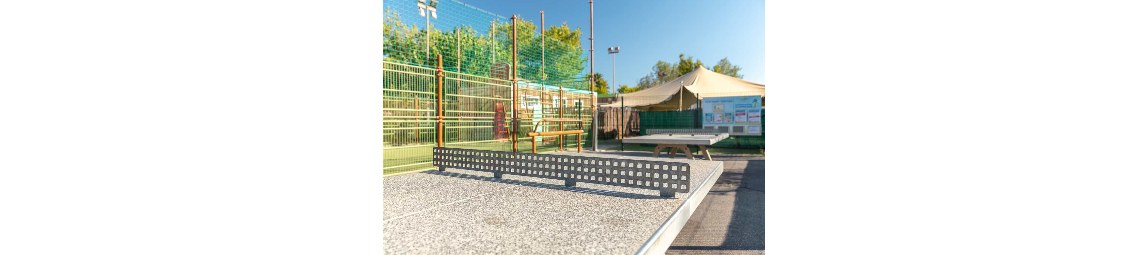 A concrete table tennis table with a perforated metal net stands in an outdoor recreational area. In the background, there is a fenced sports court, a bench, and a large canopy tent providing shade. Informational boards are visible near the tent, suggesting a community or park setting. The scene is brightly lit with clear blue skies and lush green trees, creating a vibrant and active atmosphere.