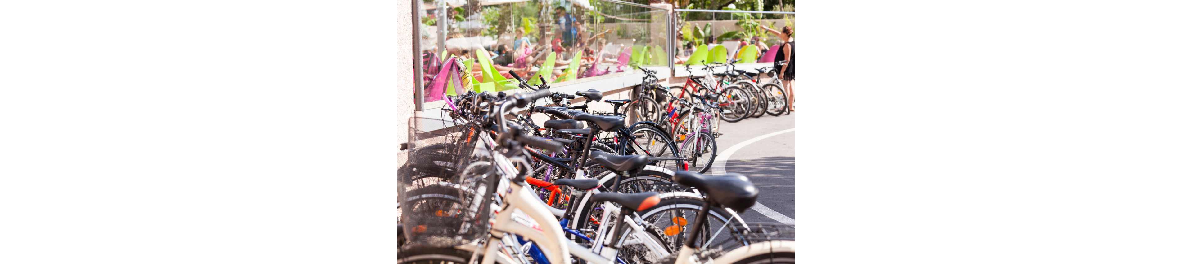 Multiple bicycles are parked closely together along a curved bike rack in an urban outdoor setting. In the background, people relax on colorful chairs behind a glass barrier, suggesting a café or public plaza. The scene is brightly lit with warm sunlight, highlighting the variety of bikes and their details. The overall atmosphere feels casual, active, and summery.