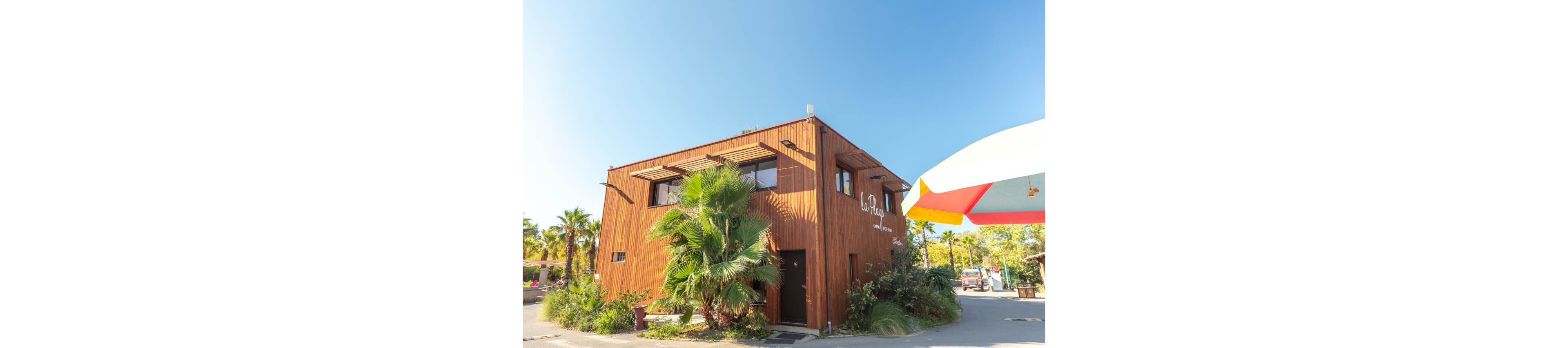A contemporary two-story wooden building stands in a sunny outdoor setting. Lush palm trees and green shrubs surround the structure, adding a tropical feel. The sky is clear and blue, suggesting a warm, bright day. Part of a colorful canopy or umbrella is visible on the right side, hinting at a nearby recreational or leisure area.