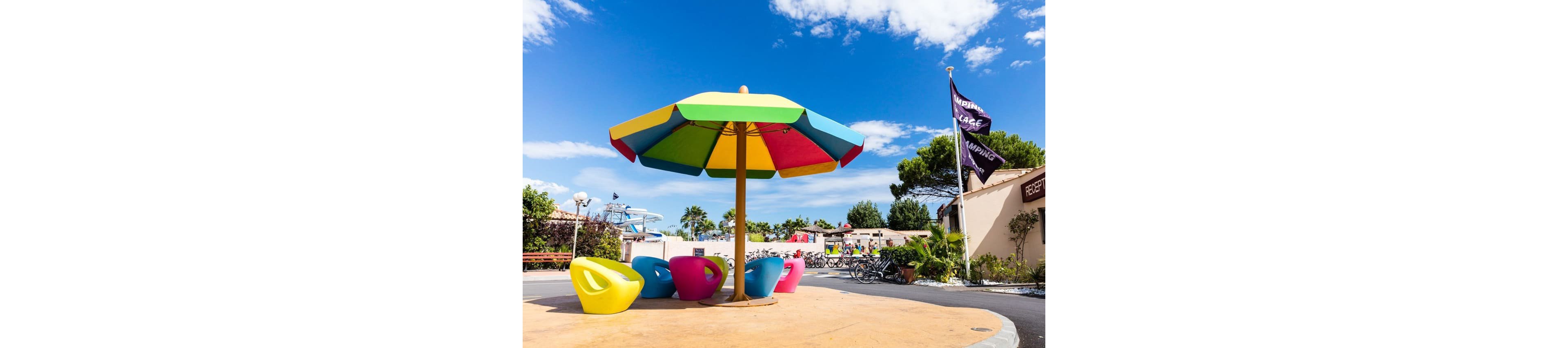A group of brightly colored modern chairs sits beneath a large multicolored umbrella in an outdoor recreational area. The scene appears to be part of a holiday campsite or resort, with bicycles, buildings, and flags visible in the background. Clear blue sky and scattered clouds create a sunny, cheerful atmosphere. The vivid colors of the furniture and umbrella contrast with the neutral ground and surrounding greenery.
