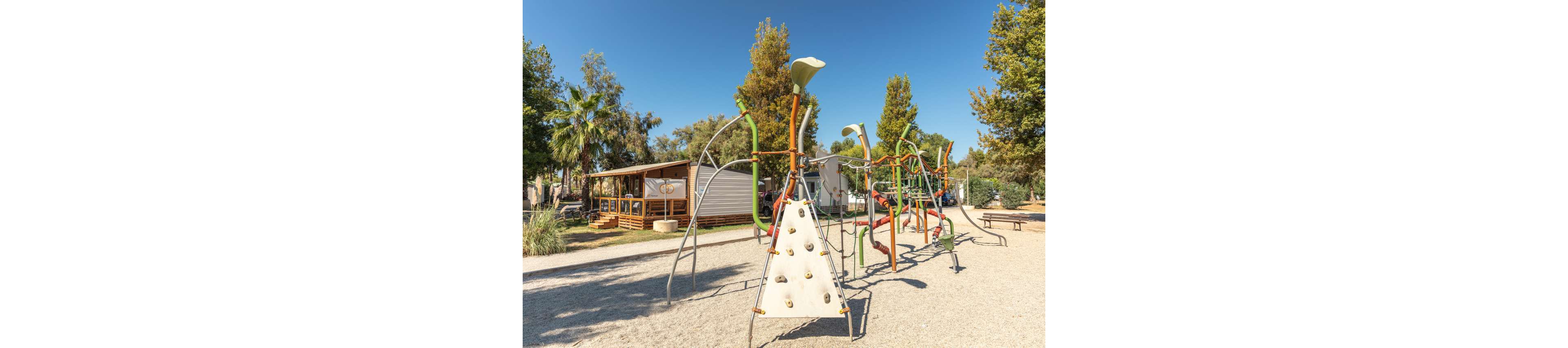 A contemporary playground with climbing structures stands in a sunny outdoor recreation area. The equipment includes a triangular climbing wall and interconnected metal frames set on a gravel surface. In the background, wooden cabins with porches and railings are surrounded by tall trees and palm trees. The bright daylight and clear blue sky create a warm, inviting atmosphere suitable for families and children.