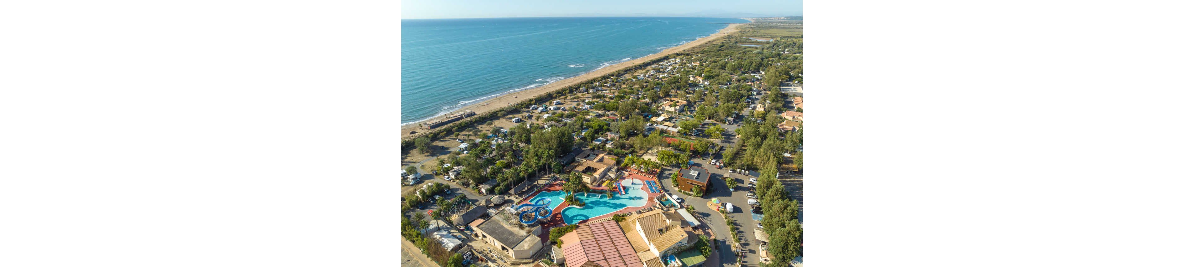 A large coastal campsite with a central waterpark is captured from an aerial perspective. The scene shows multiple swimming pools, water slides, and surrounding buildings nestled among trees. Numerous caravans, tents, and small structures are arranged in neat rows extending toward the shoreline. A long sandy beach and calm sea stretch along the left side of the image, emphasizing the seaside location. The overall atmosphere suggests a busy yet relaxed holiday resort environment.