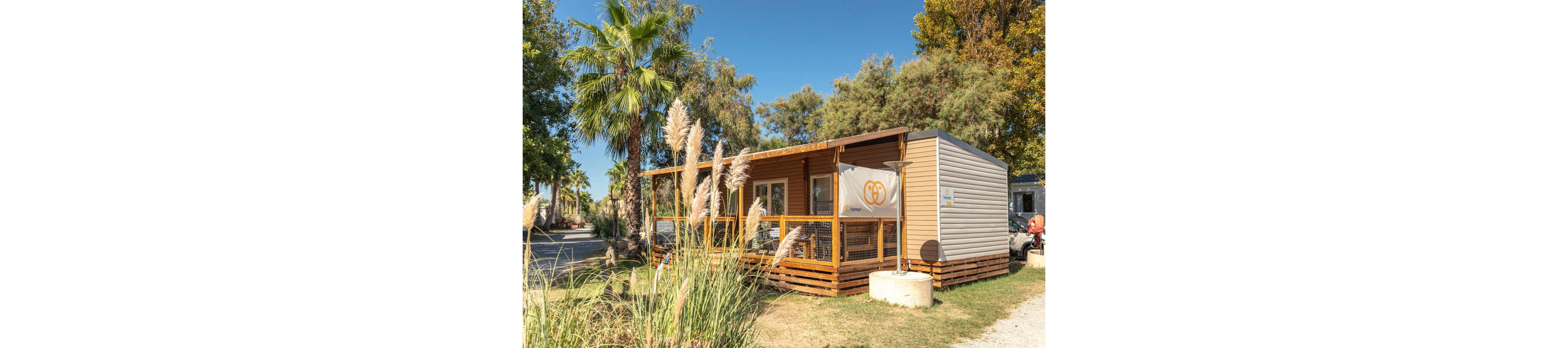 A modern mobile home with a wooden deck stands in a sunny campsite surrounded by trees and ornamental grasses. The structure features light-colored siding, large windows, and a covered porch with railing. A palm tree and other lush greenery frame the scene, suggesting a warm climate. The clear blue sky and bright daylight create a relaxed vacation atmosphere.