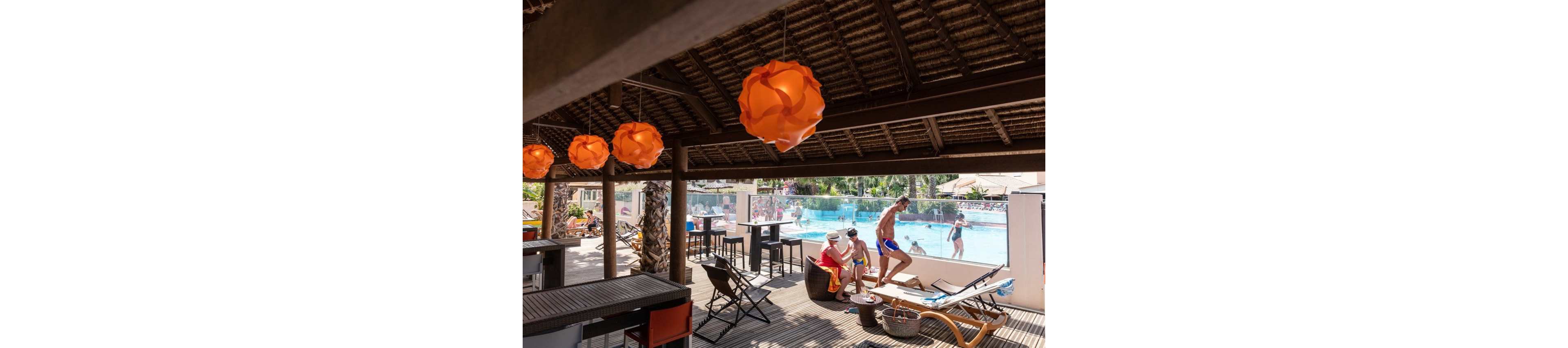 An open-air resort bar overlooks a lively swimming pool where guests enjoy a sunny day. Orange pendant lights hang from the thatched roof, casting warm tones over the seating area. People sit and chat on lounge chairs while others play and swim in the pool in the background. The scene conveys a relaxed vacation atmosphere with casual summer clothing and bright colors.