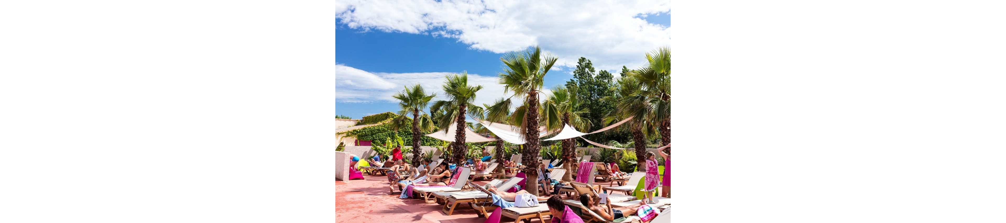 A group of people lounge on sunbeds under tall palm trees at an outdoor poolside area. Colorful towels and swimsuits add vibrancy to the scene as guests relax and sunbathe. White shade sails provide partial cover from the bright sun, while a blue sky with scattered clouds stretches overhead. The setting feels like a tropical resort or vacation destination with a casual, leisurely atmosphere.
