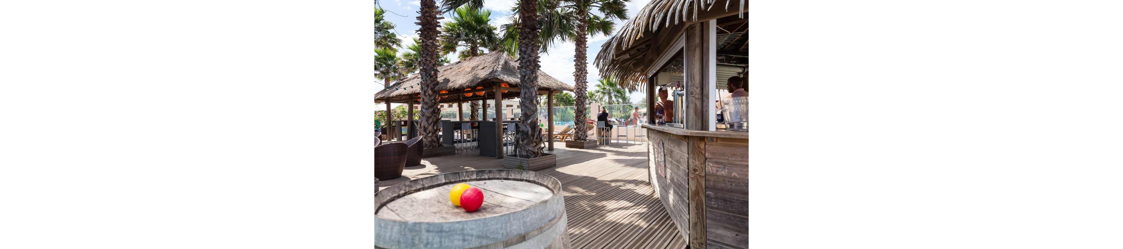 An outdoor tropical bar area features wooden decks lined with tall palm trees and thatched-roof huts. A rustic barrel in the foreground holds two colorful balls, adding a playful touch to the scene. People relax and socialize around the bar and seating areas in the background. Bright daylight and clear skies create a warm, summery atmosphere.