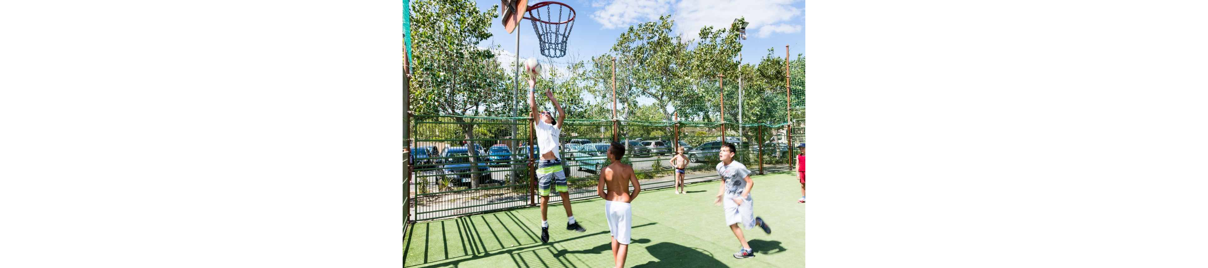 A group of boys play an energetic game of basketball on an outdoor court. One player jumps high to shoot the ball toward the hoop while others watch and prepare to react. The court is enclosed by a metal fence with parked cars and trees visible in the background. Bright daylight and clear skies create strong shadows on the green playing surface, emphasizing the lively, active atmosphere.