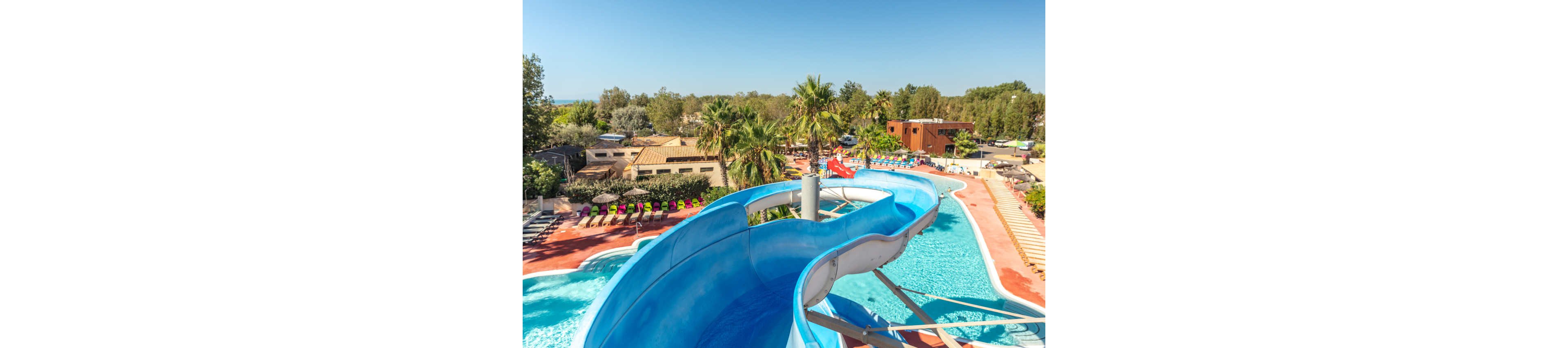 A bright blue waterslide curves into a large outdoor swimming pool at a sunny resort. Lounge chairs and umbrellas line the poolside area, creating a relaxed vacation atmosphere. Palm trees and low buildings surround the waterpark, with additional pools and slides visible in the background. The clear sky and vibrant colors emphasize a warm, family-friendly holiday setting.