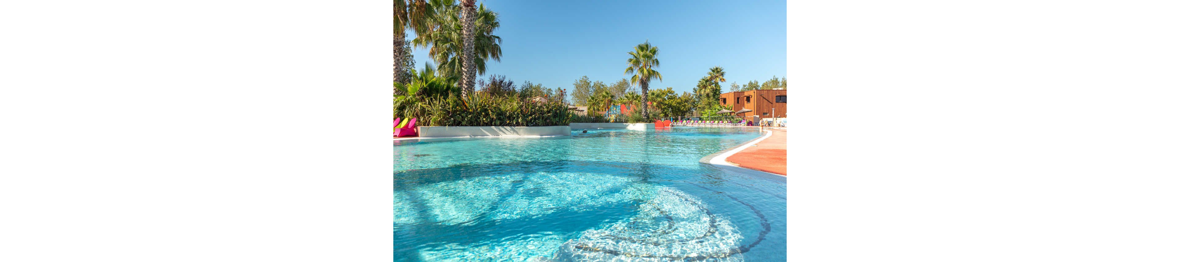 A clear blue swimming pool glistens under bright sunlight at a resort. Palm trees and lush greenery surround the water, creating a tropical atmosphere. Colorful lounge chairs and umbrellas line the far side of the pool near modern low-rise buildings. The scene feels calm and inviting, ideal for relaxation and leisure. The sky is clear and blue, enhancing the vibrant, summery mood.
