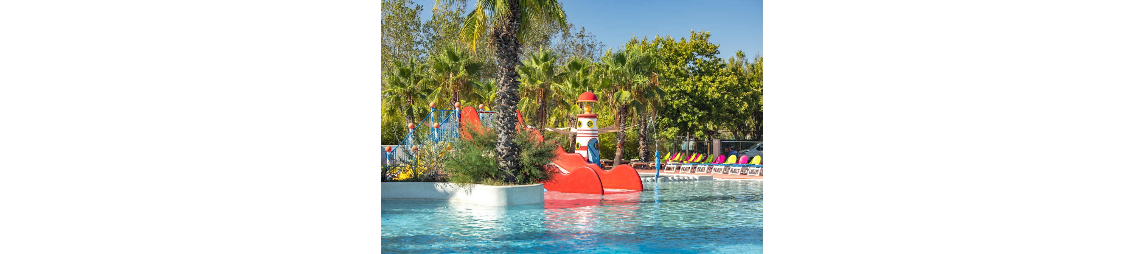 A vibrant children’s water playground features bright red slides and a whimsical lighthouse structure in a shallow pool. Palm trees and lush greenery surround the area, creating a tropical resort atmosphere. In the background, rows of colorful lounge chairs line the poolside, suggesting a family-friendly vacation setting. The clear blue water and sunny sky emphasize a fun and relaxing outdoor environment.