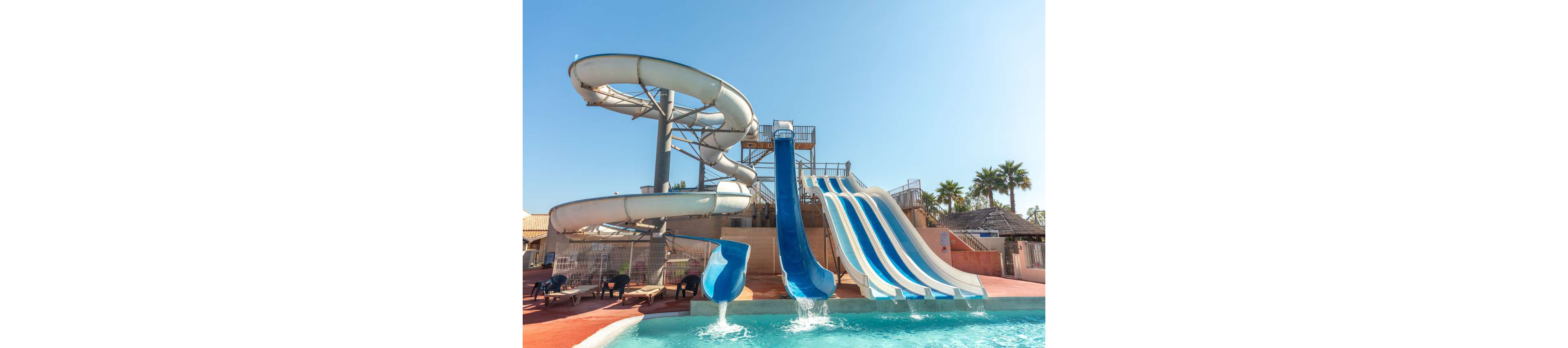A large outdoor water park features several bright blue and white slides emptying into a clear swimming pool. A tall spiral tube slide winds down from a metal platform beside a set of open straight slides. The scene is set under a clear blue sky with palm trees and resort-style buildings in the background. Sun loungers and shaded areas line the pool deck, suggesting a family-friendly vacation atmosphere.