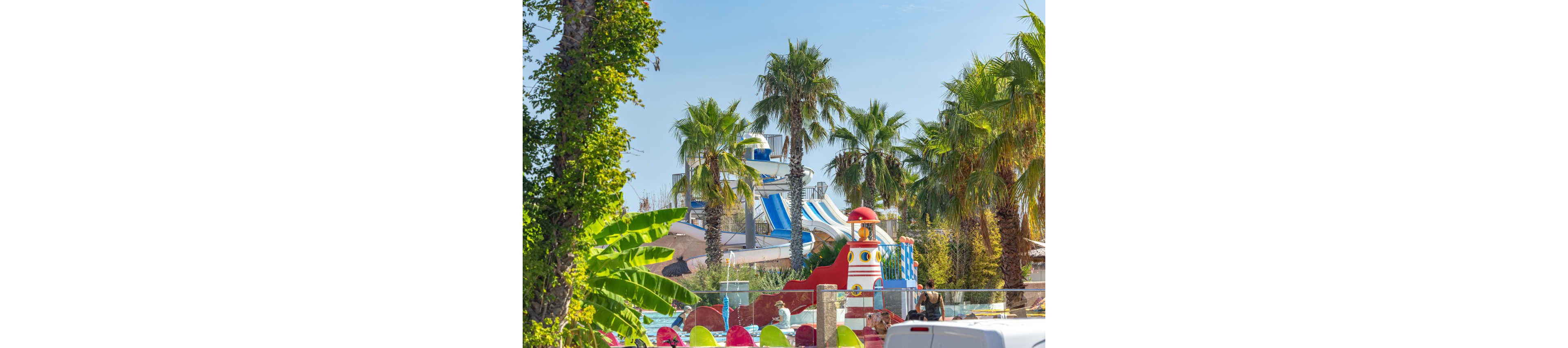 A vibrant water park is set among tall palm trees under a clear blue sky. Large striped water slides and a playful, ship-themed structure dominate the background. In the foreground, bright inflatable tubes and colorful features add to the lively atmosphere. The scene suggests a fun, family-friendly destination in a warm, tropical climate.