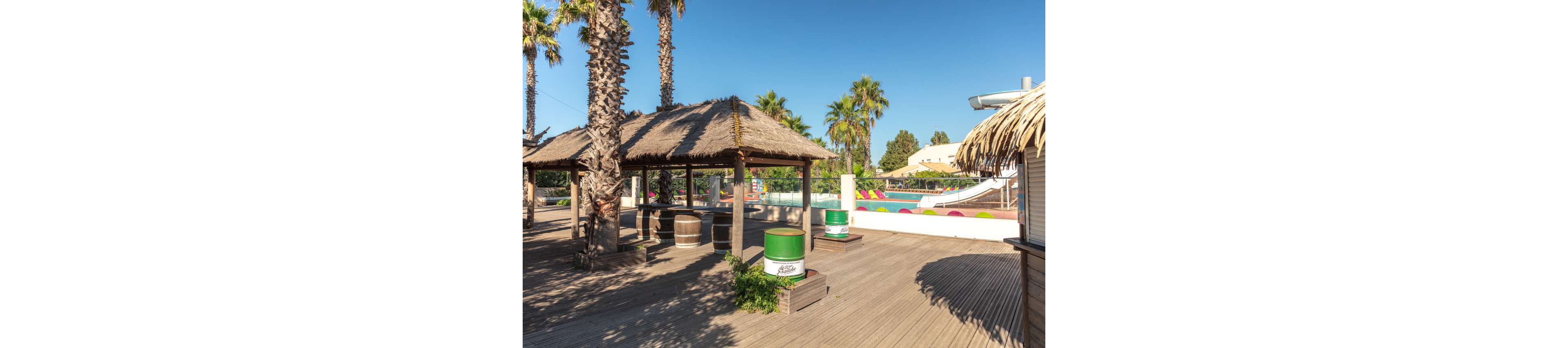 An outdoor tropical-style bar area is set on a wooden deck surrounded by tall palm trees. Thatched-roof huts provide shaded seating near a bright swimming pool with colorful lounge chairs and slides in the background. Green and white barrel tables and a small hut structure add to the resort-like atmosphere. The clear blue sky and strong sunlight create a warm, summery vacation setting.