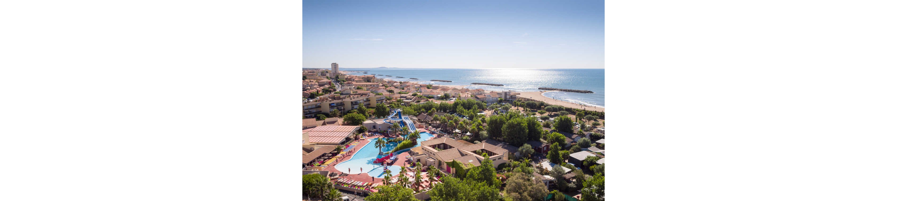 An aerial view shows a large coastal resort featuring a bright blue swimming pool and winding water slides. The complex is surrounded by lush greenery, low-rise buildings, and sun loungers arranged around the pool. Beyond the resort, a sandy beach stretches along the shoreline with calm sea waters and breakwaters visible offshore. The clear sky and expansive ocean create a relaxed vacation atmosphere in this seaside town.