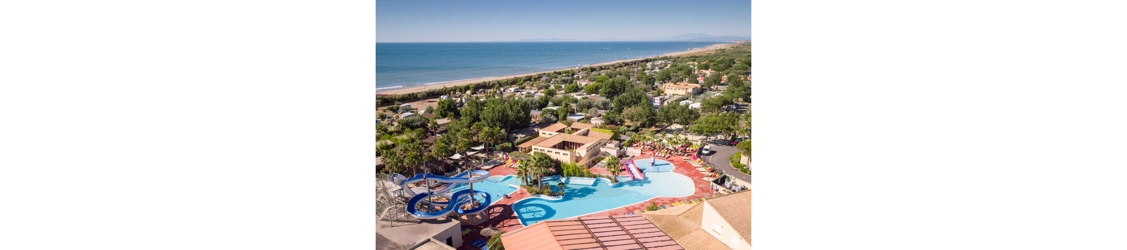 An aerial view shows a coastal holiday resort featuring a large outdoor swimming pool complex with winding blue water slides. The pool area is surrounded by sun loungers, palm trees, and low-rise buildings that appear to house resort facilities. Beyond the resort, a campsite with numerous plots and trees stretches toward a long sandy beach. The calm sea and distant coastline create a relaxed vacation atmosphere under clear skies.