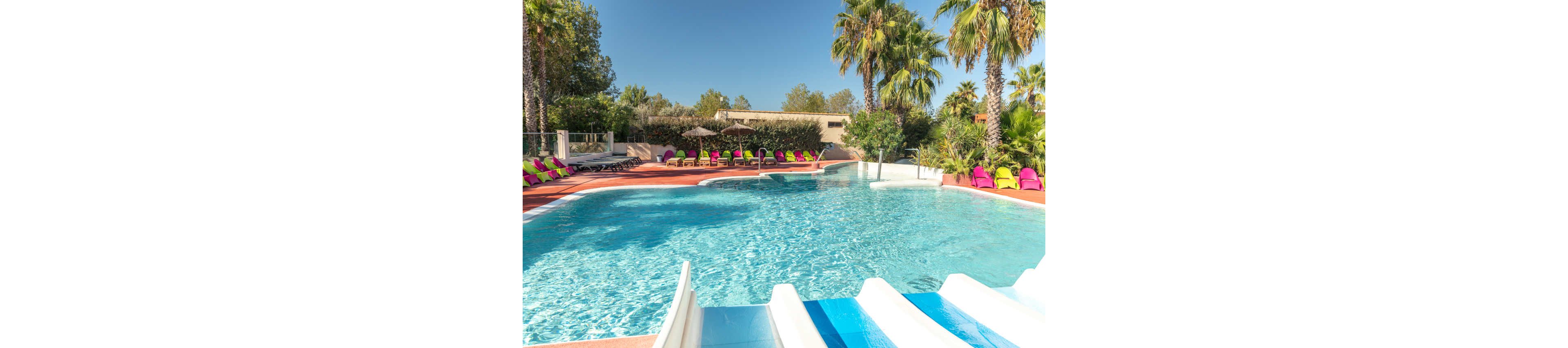 A bright outdoor swimming pool is shown under a clear sky, featuring a white and blue waterslide in the foreground. Colorful lounge chairs line the poolside deck, arranged neatly around the water. Tall palm trees and lush greenery surround the area, creating a tropical resort atmosphere. The scene suggests a relaxing vacation setting ideal for families and leisure.