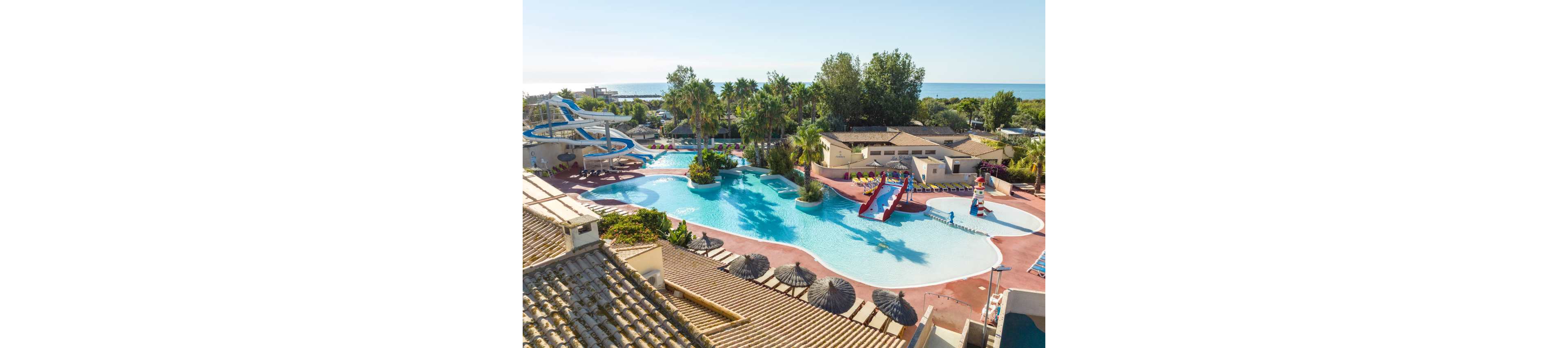 An aerial view shows a large resort swimming pool complex with multiple curving waterslides and shallow play areas. The pool is surrounded by palm trees, sun loungers, and low-rise buildings with tiled roofs. Bright red and blue slides lead into the water, creating a playful family-friendly atmosphere. In the background, the sea and coastline are visible beyond the resort, emphasizing its coastal holiday setting.