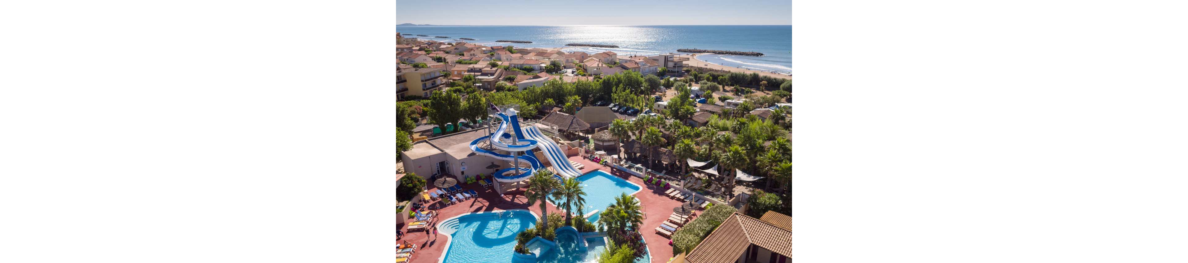 A seaside resort features a large swimming pool complex with multiple blue and white waterslides. Sun loungers and umbrellas surround the pools, where guests relax in the warm weather. The resort is nestled among trees and low-rise buildings, with a sandy beach and calm sea in the background. The bright sunlight and clear sky create a vibrant, summery atmosphere overlooking the coastline.