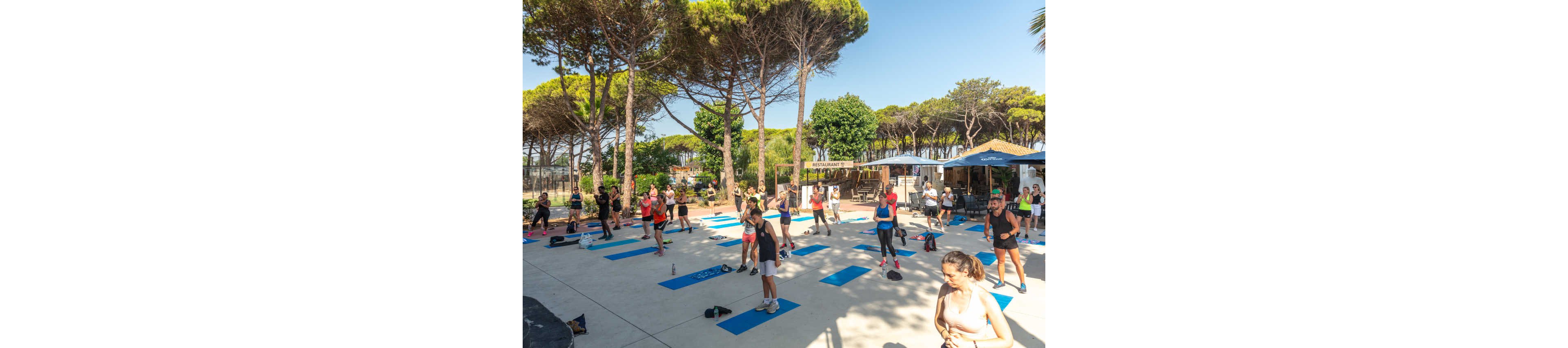 A large group of people participate in an outdoor fitness class on a sunny day. Exercise mats are spaced out on a paved area surrounded by tall pine trees. Participants wear athletic clothing and appear to be following an instructor in the center. In the background, a small café or bar area with umbrellas and a restaurant sign is visible, along with other onlookers.