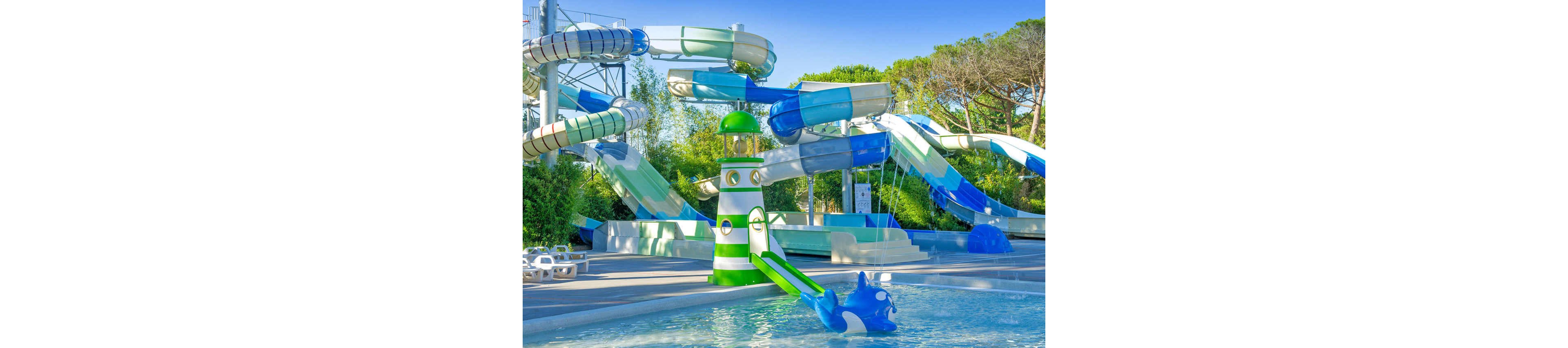 An outdoor water park features multiple large twisting water slides in shades of blue, green, and white. In the foreground, a shallow pool includes a playful lighthouse-themed slide and a blue animal-shaped water toy. Surrounding the pools are sun loungers and trees, creating a relaxed holiday atmosphere. The scene is brightly lit under a clear blue sky, emphasizing a fun and family-friendly environment.