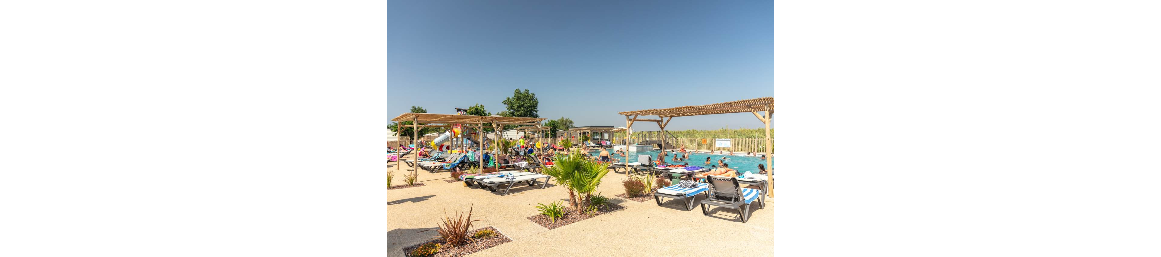 An outdoor swimming pool is filled with people relaxing and playing in the water at a sunny resort. Guests lounge on sunbeds under wooden pergolas around the pool deck. A waterslide and play area are visible in the background, adding to the family-friendly atmosphere. The sky is clear and blue, and small palm plants and landscaped beds decorate the foreground.