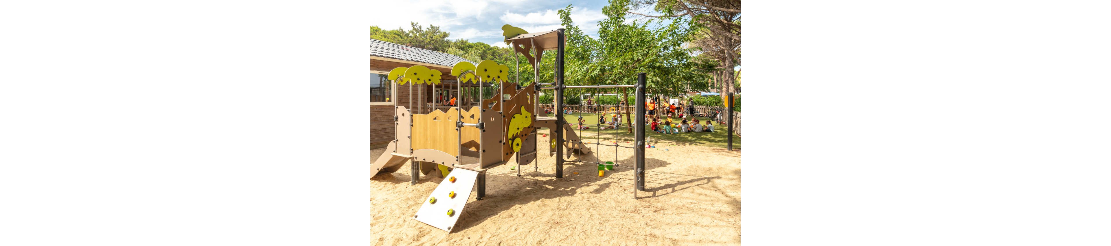 A modern wooden playground structure stands on a sandy surface in a sunny outdoor area. The play equipment includes climbing holds, a slide, and rope elements surrounded by trees and greenery. In the background, a group of children and adults gather on the grass near a building. The scene feels bright and cheerful, suggesting an active day at a park or schoolyard.