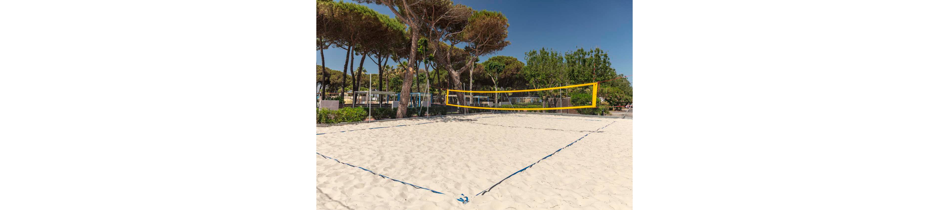 An empty sand volleyball court is set up outdoors under a clear blue sky. The court features a bright yellow net and blue boundary lines on clean white sand. Tall pine trees and green shrubs surround the area, providing a natural backdrop. Buildings and seating areas are visible in the distance, suggesting a recreational or resort setting.