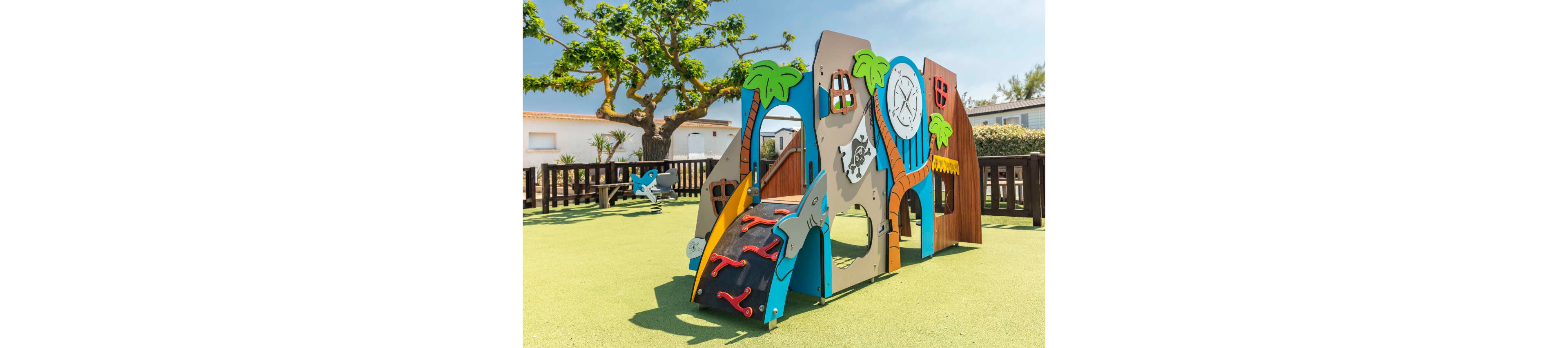 A brightly colored pirate-themed play structure stands in an outdoor playground. The equipment features a small climbing wall, slide, and decorative elements like palm trees, a treasure map, and a pirate flag. It is set on a soft green safety surface, surrounded by a wooden fence and nearby houses. The scene is sunlit with clear skies and a large tree providing additional visual interest.