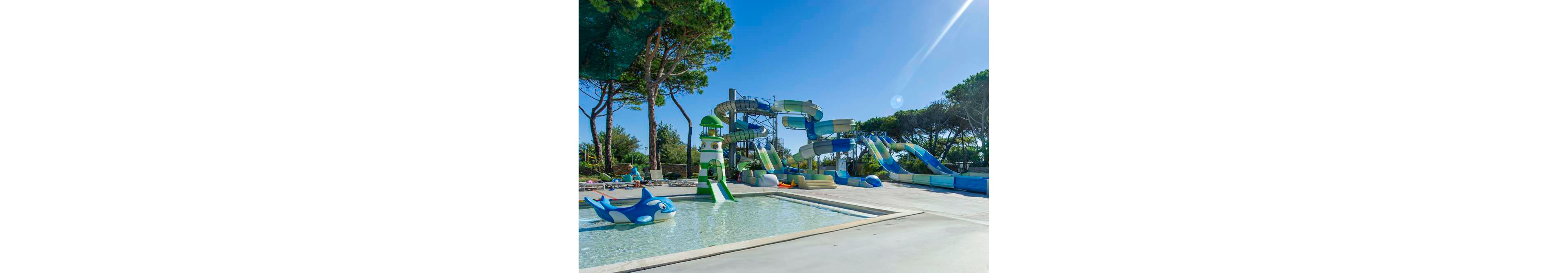 A group of people lounge on sunbeds beside a shallow outdoor pool at a waterpark. Large twisting water slides and play structures rise in the background under a clear blue sky. Tall pine trees surround the area, creating a natural, shaded backdrop. Bright swimwear, inflatable toys, and sparkling water convey a fun, summery vacation atmosphere.
