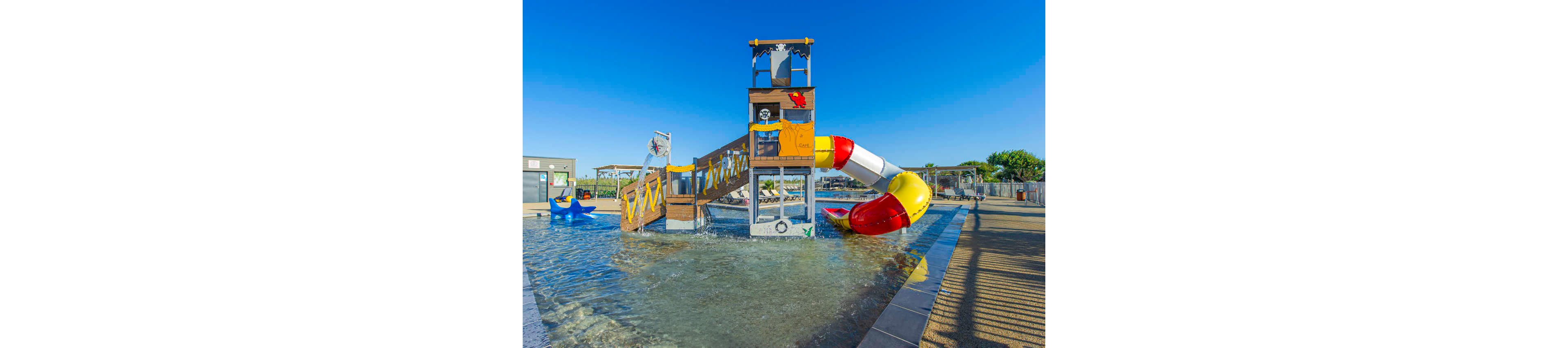 A group of adults and children play together in a shallow outdoor waterpark pool. The setting features a colorful pirate-themed play structure with slides, flags, and water features under a clear blue sky. People splash, walk, and run through the water, creating a lively and energetic scene. The bright sunlight and vivid colors give the image a cheerful, summery atmosphere.