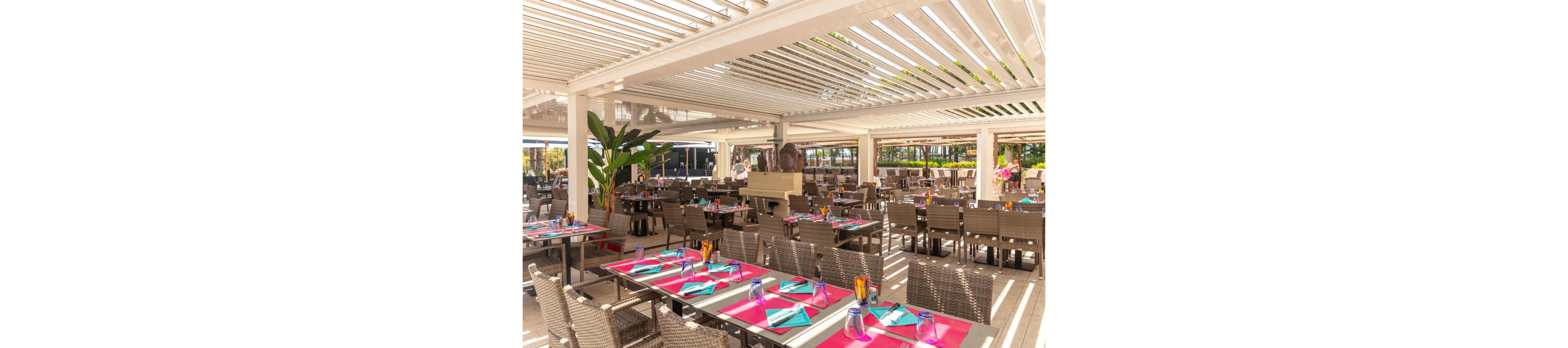 An open-air restaurant terrace is set with numerous wicker chairs and neatly arranged tables. Bright pink placemats and turquoise napkins add vibrant color under a white slatted pergola roof. Sunlight filters through the overhead structure, creating striped shadows across the floor and furniture. The space appears ready for guests, with glassware, cutlery, and decorative plants enhancing the inviting atmosphere.