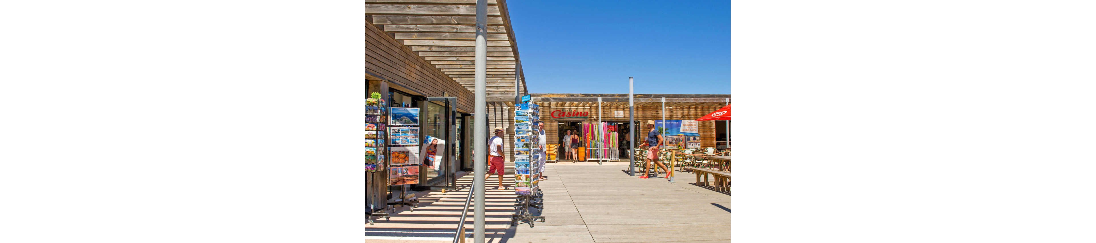 An outdoor boardwalk lined with wooden seaside shops is shown under a bright blue sky. Several postcard racks stand along the walkway, displaying colorful images for tourists. People in casual summer clothing browse the displays and walk between the stores. A café terrace with wooden tables and umbrellas is visible on the right, enhancing the relaxed holiday atmosphere.