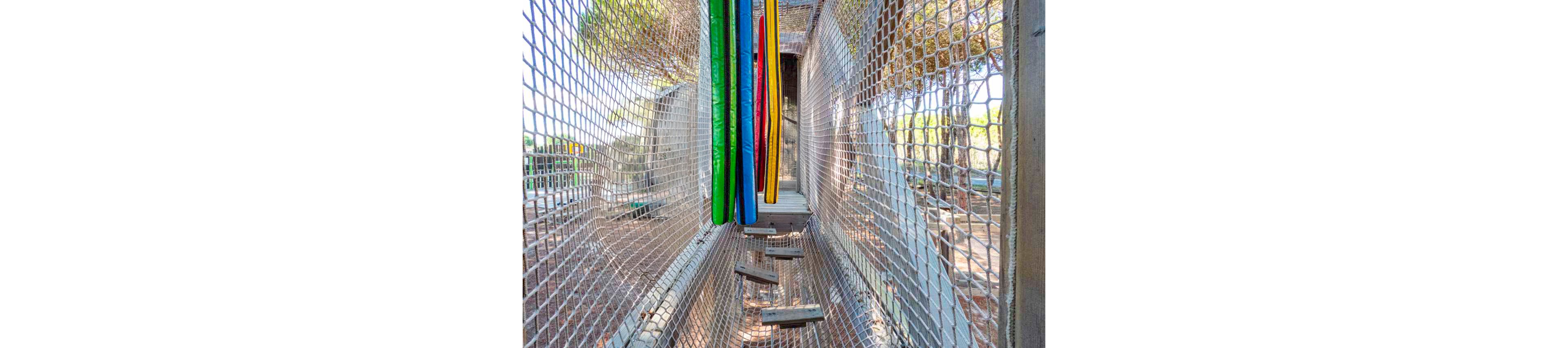 Two children play together on a suspended rope bridge in an outdoor adventure park. They balance on wooden planks while holding onto bright, multicolored padded poles. The structure is enclosed by safety netting, with trees and sunlight visible in the background. The scene feels energetic, playful, and summery.