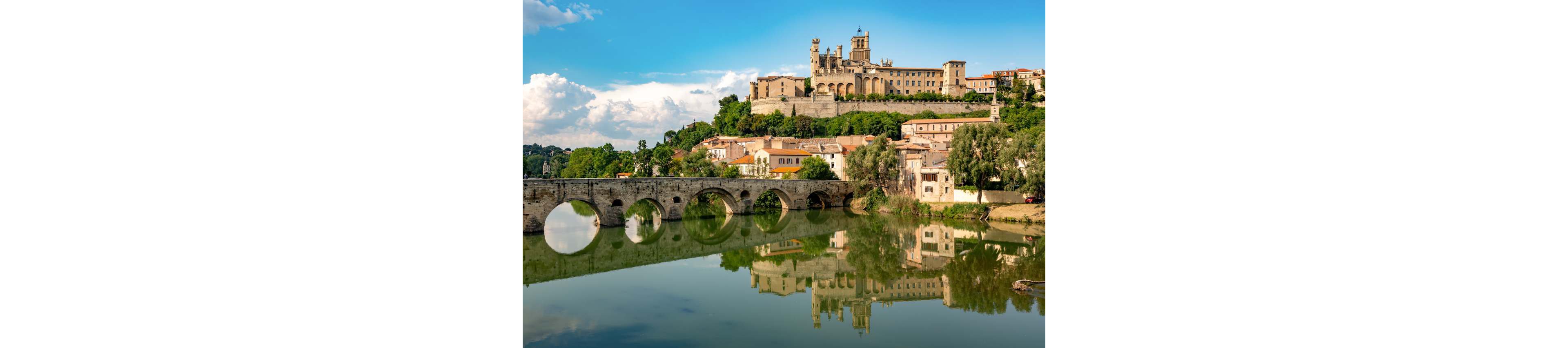 A historic hilltop town with a large cathedral overlooks a calm river. An old stone arch bridge spans the water, its arches reflected clearly in the surface below. Traditional houses with tiled roofs cluster along the riverbank beneath the imposing church and fortifications. Lush green trees and a partly cloudy blue sky complete the picturesque landscape.
