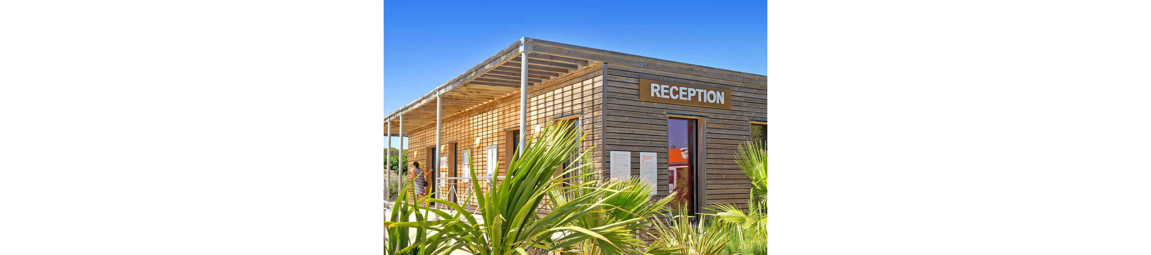 A contemporary wooden reception building stands under a clear blue sky. The structure features horizontal slats, a flat pergola-style roof, and large windows reflecting the bright daylight. A prominent sign reading "RECEPTION" is mounted above the entrance. Green tropical plants frame the foreground, and a person is visible near the doorway, suggesting an active visitor area.