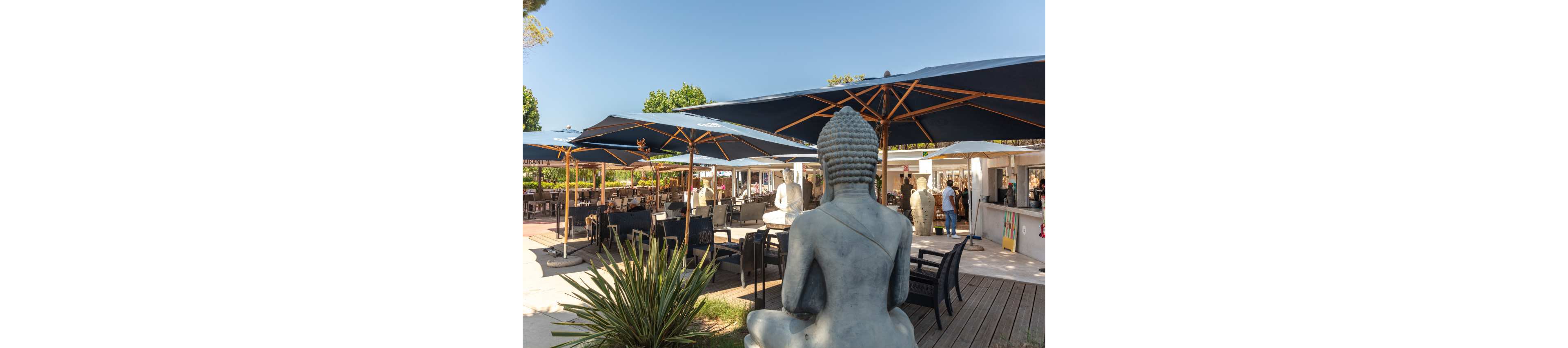 A large Buddha statue overlooks an open-air café terrace. Dark sun umbrellas shade seating areas with tables and lounge chairs on wooden decking. People are gathered near the bar and walking through the bright, sunny space. Green plants and trees frame the relaxed outdoor setting under a clear blue sky.