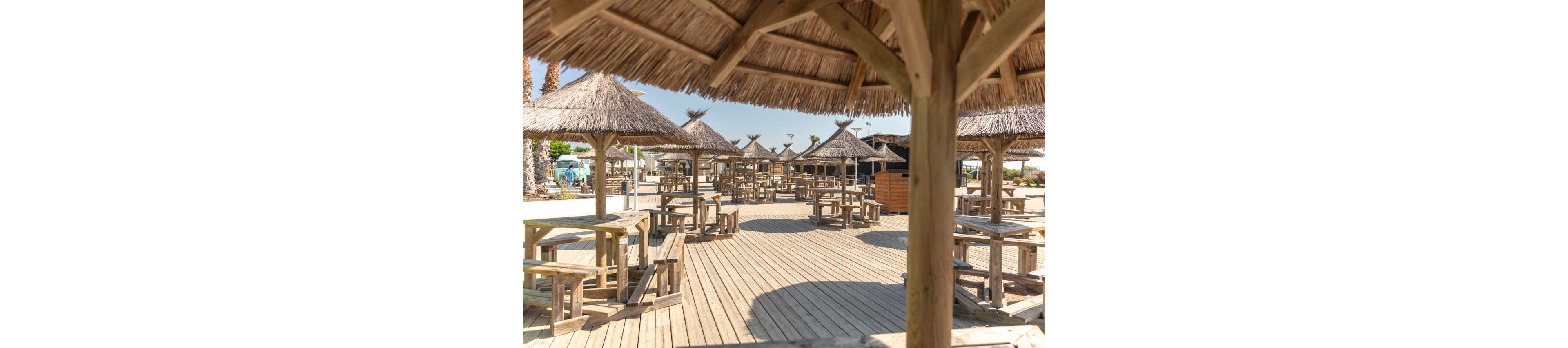 A spacious outdoor terrace features numerous wooden picnic tables under straw-thatched umbrellas. The area is built on a wide wooden deck, suggesting a beachside or coastal setting. Bright sunlight and clear skies create a warm, summery atmosphere. No people are present, emphasizing the calm and open space ready for visitors.