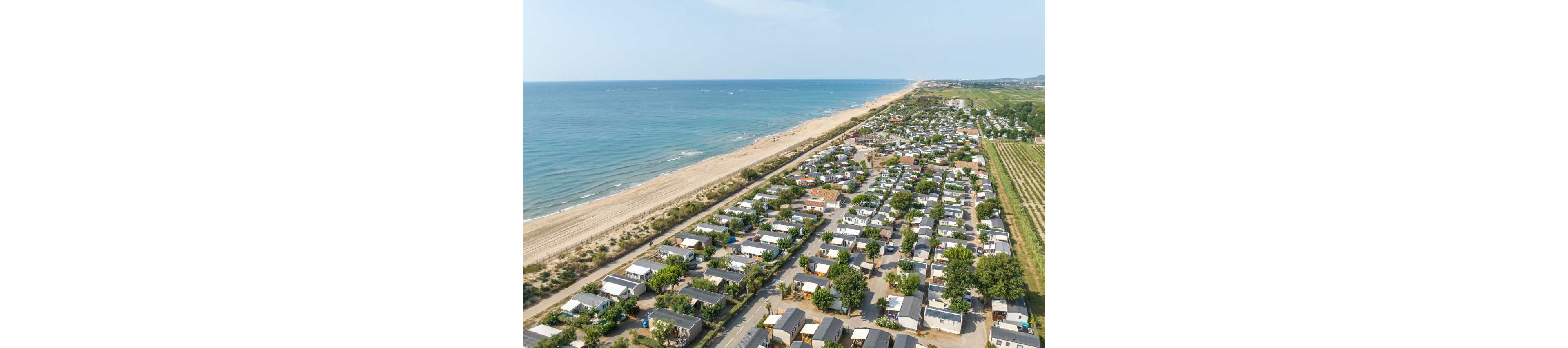 A wide aerial view shows a large campground or holiday park situated along a sandy coastline. Rows of small cabins, mobile homes, and RV-style units are arranged in an orderly grid with trees scattered between them. The expansive beach and calm sea stretch along one side, while agricultural fields or countryside extend into the distance on the other. The scene suggests a relaxed vacation destination with easy access to the ocean and outdoor recreation.