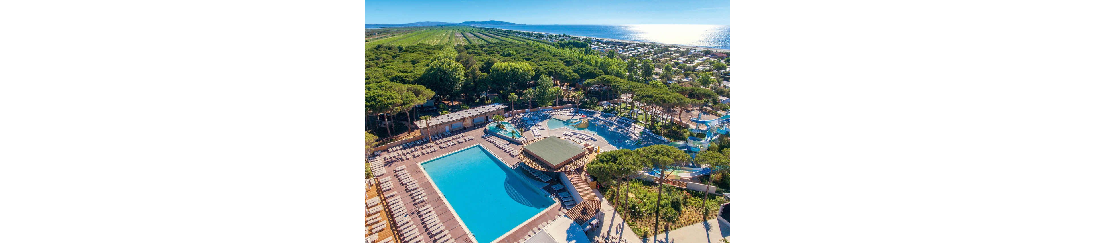A large holiday resort with multiple swimming pools is captured from an aerial perspective near a coastline. The main rectangular pool is surrounded by rows of sun loungers and shaded seating areas. Additional circular pools and water slides are visible among tall pine trees, creating a family-friendly water park atmosphere. In the background, the resort extends toward the sea, with campsites or bungalows and agricultural fields stretching into the distance under clear blue skies.