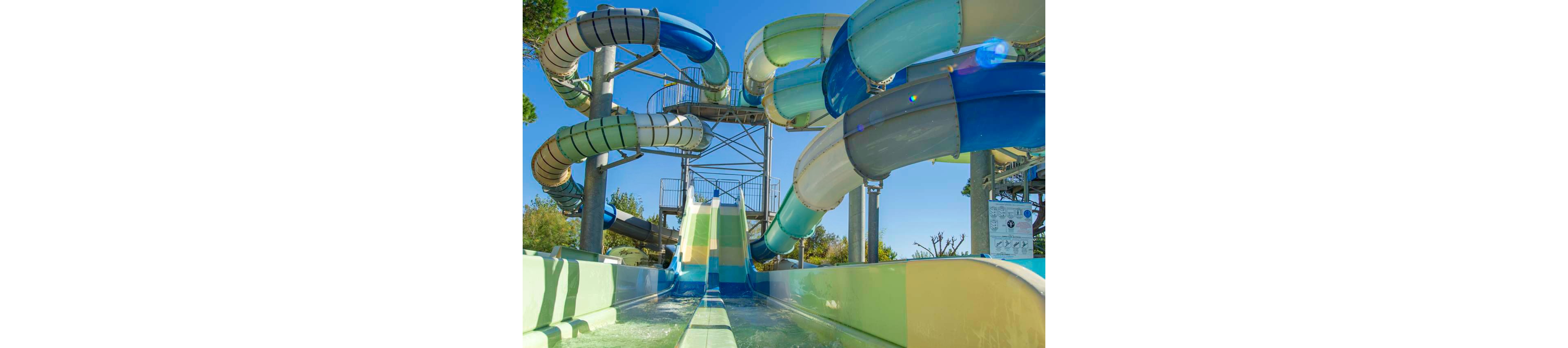 Two young women in bright swimsuits walk through shallow water at the end of a large water slide. They smile and high-five each other, suggesting a fun and carefree summer outing. Behind them, multiple twisting water slides and support structures dominate the scene under a clear blue sky. The vibrant colors and splashing water create an energetic, playful atmosphere in an outdoor water park.