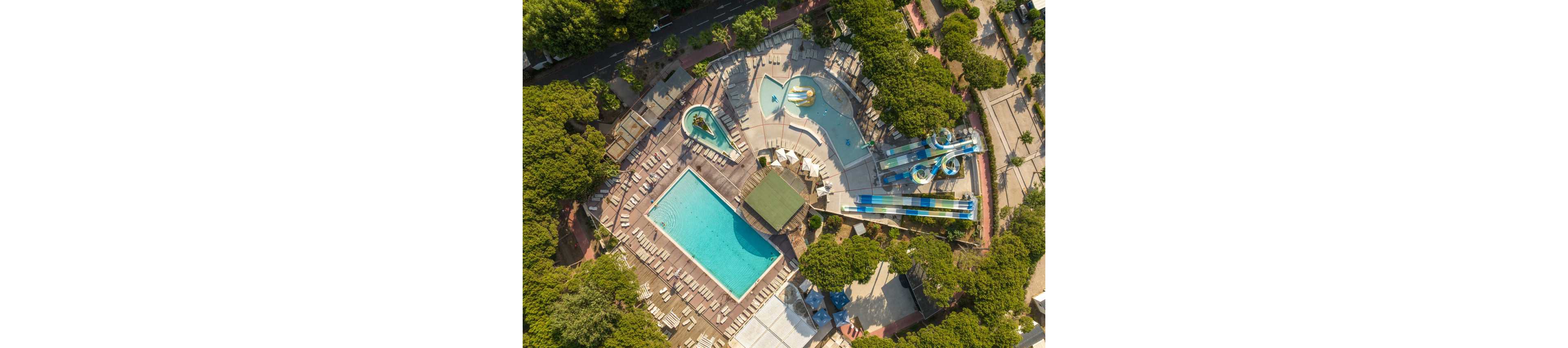 A large outdoor water park is photographed from above, showing multiple pools and colorful water slides. A rectangular swimming pool sits beside a curving leisure pool with play structures and splash areas. Numerous empty lounge chairs surround the pools, indicating a quiet time with no visitors present. Dense green trees frame the complex, and nearby walkways and buildings outline the park’s layout.