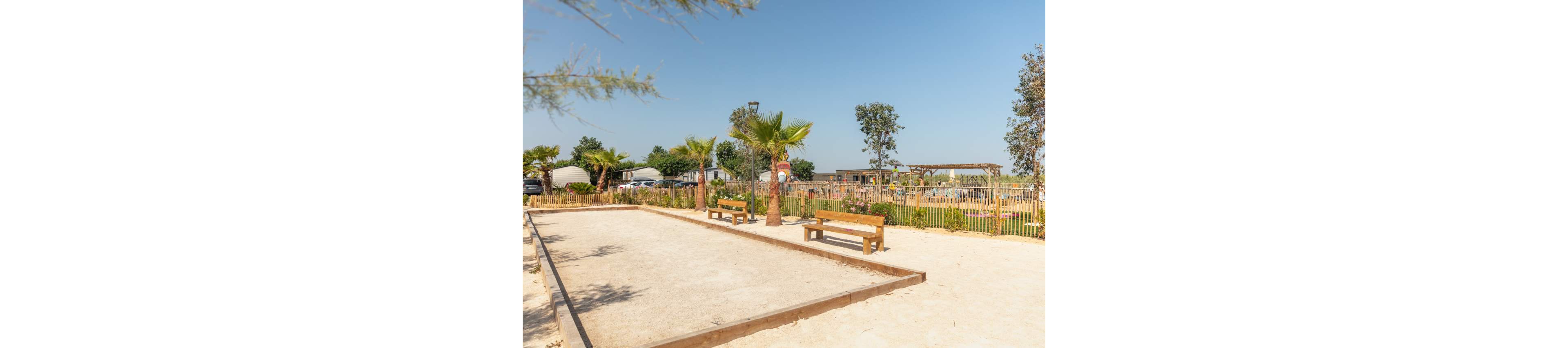 An empty outdoor bocce or pétanque court sits under a clear blue sky in a landscaped holiday park. Wooden benches line the sandy playing area, offering seating for players and spectators. Palm trees, shrubs, and a wooden fence frame the court, adding a relaxed, resort-like atmosphere. In the background, small buildings and a pergola structure suggest campsite or vacation accommodations.