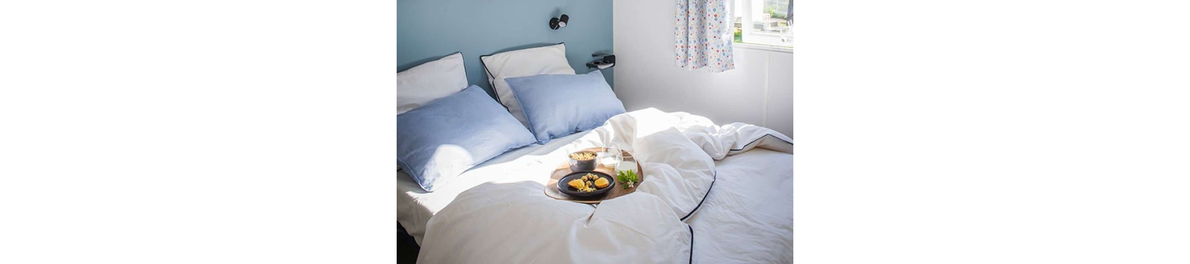 A neatly made double bed with white linens and light blue pillows is set in a bright, sunlit bedroom. A wooden tray with breakfast items, including a bowl of cereal, a glass of milk or water, and a small plate of food, rests on the rumpled duvet. Soft daylight streams in through a nearby window with patterned curtains, illuminating the scene. The overall atmosphere feels calm, inviting, and leisurely, suggesting a relaxed morning at home.